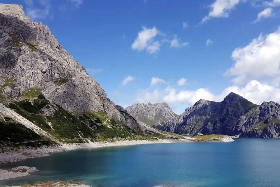 Turquoise alpine lake sits below rocky gray mountains with a pale shoreline curving into the distance beneath a partly cloudy sky. Remote landscape often found along hiking trails in Europe.