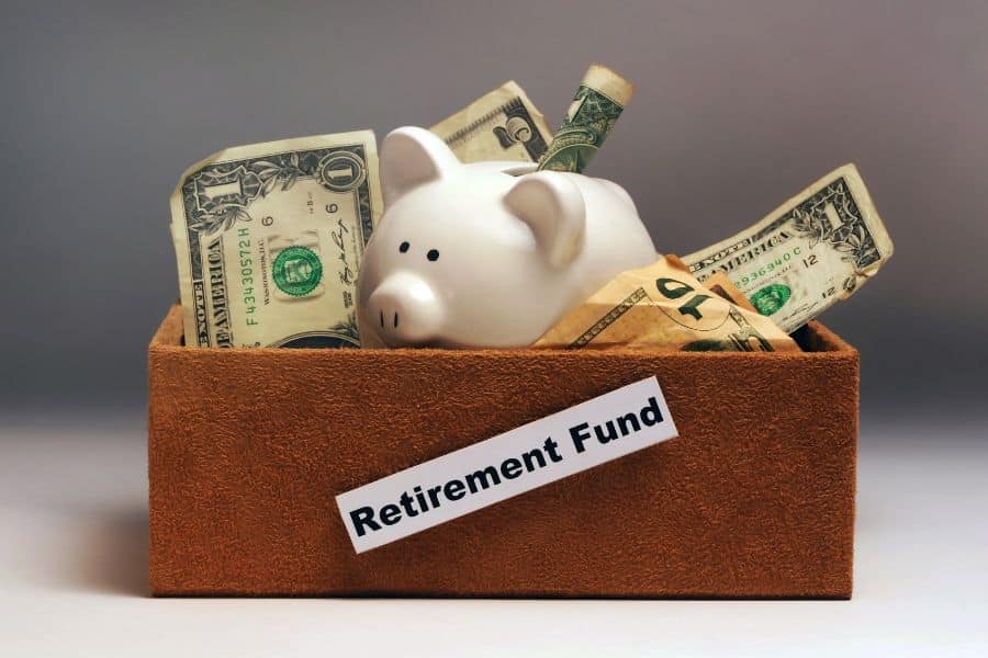 White piggy bank filled with dollar bills sits inside a brown box labeled "Retirement Fund." The image represents saving money over time for retirement.