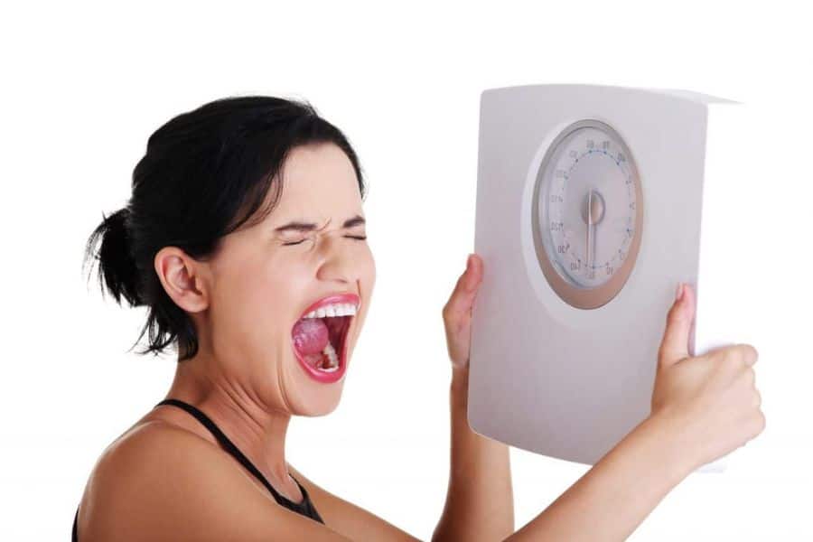 Woman with eyes shut and mouth open in frustration while holding a bathroom scale against a plain white background. The photo shows the emotional stress that can come with tracking body weight.