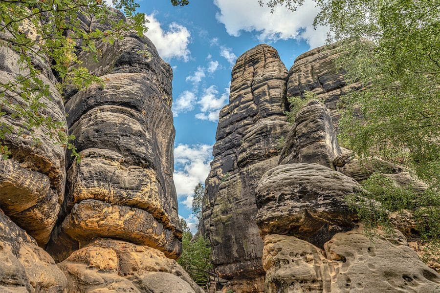 Tall weathered sandstone rock towers rise above a forested path, with textured cliffs framing a slice of blue sky and clouds. Rugged rock formations beside hiking trails in Europe.