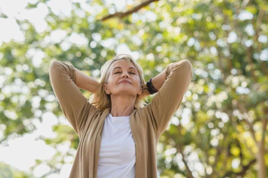 Older woman stands beneath leafy trees with her hands behind her head and her eyes closed in a relaxed pose. The photo conveys a peaceful pause in nature.