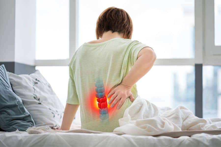 Person sitting upright in bed with one hand pressed against the lower back while a glowing red spine graphic marks the painful area. This sciatica themed illustration emphasizes lower back pain radiating from the lumbar spine.