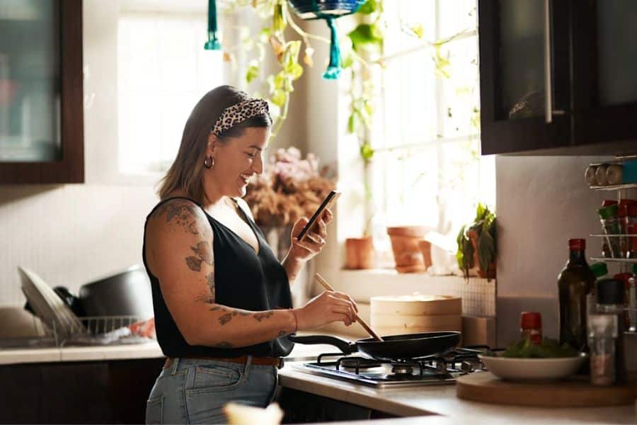 Woman smiling at her phone while cooking at the stove in a sunlit kitchen with plants hanging near the window. The photo suggests following recipes or tracking meals as part of healthier daily habits.