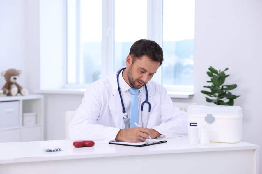 Doctor in a white coat sits at a desk writing notes during a consultation about hand grip test results or general health. A stethoscope, pill bottles, and a white medical kit sit on the desk in a bright exam room.