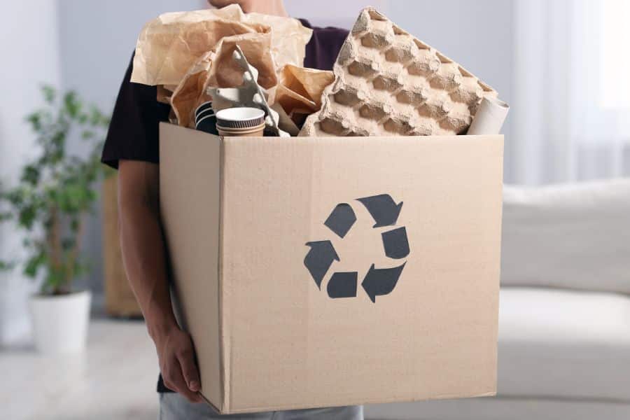 A person carries a cardboard box marked with a recycling symbol and filled with paper packaging, cups, and molded paper trays. This guide to composting suggests collecting compostable paper based materials separately from household waste when local rules allow it.