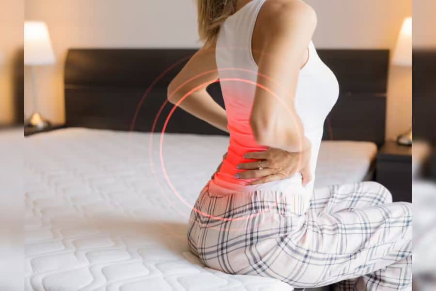 Woman sitting on the edge of a bed in plaid pajama pants while holding her lower back in pain. A red glowing highlight over her lower spine emphasizes discomfort that can be associated with joint stiffness.