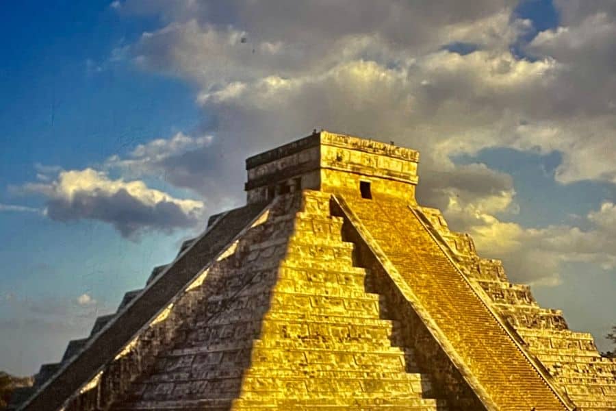 The stone pyramid at Chichen Itza rises sharply against a sky filled with large sunlit clouds. Warm golden light on the steps gives the ancient structure a dramatic look often associated with spring equinox events at the site.