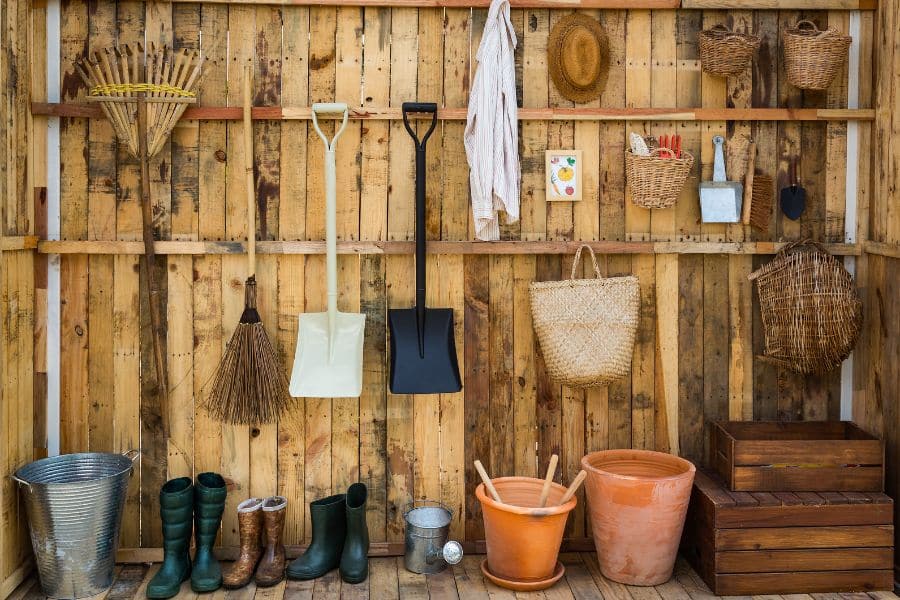 Garden tools and supplies hang neatly on a wooden shed wall, including rakes, shovels, baskets, boots, pots, and a watering can. The setup shows the basic storage and equipment that help you start a vegetable garden and keep it maintained.