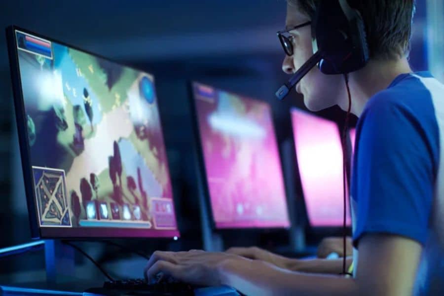 Young person wearing a gaming headset plays at a desktop computer with glowing monitors in a dark room. This supports work from home jobs content about game streaming esports or other online tech based work done from home.