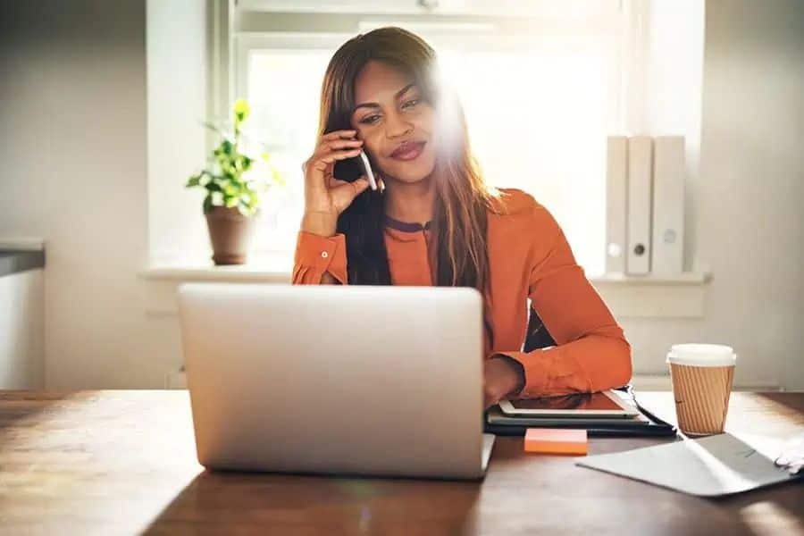 Woman talks on her phone while sitting at a table with a laptop coffee cup and office supplies near a sunny window. This is a natural fit for work from home jobs content about remote business roles freelance work or virtual meetings.