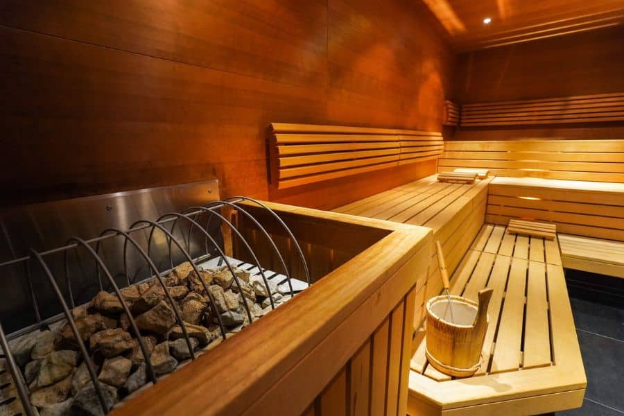 Empty wooden sauna with tiered benches warm lighting and a heater filled with stones in the foreground. The space suggests rest recovery and intentional wellness time.