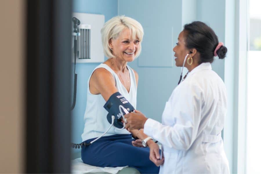 Older patient sits on an exam table smiling while a doctor checks her blood pressure in a clinic room. The photo adds context around medical evaluation and monitoring for conditions linked to stroke symptoms.
