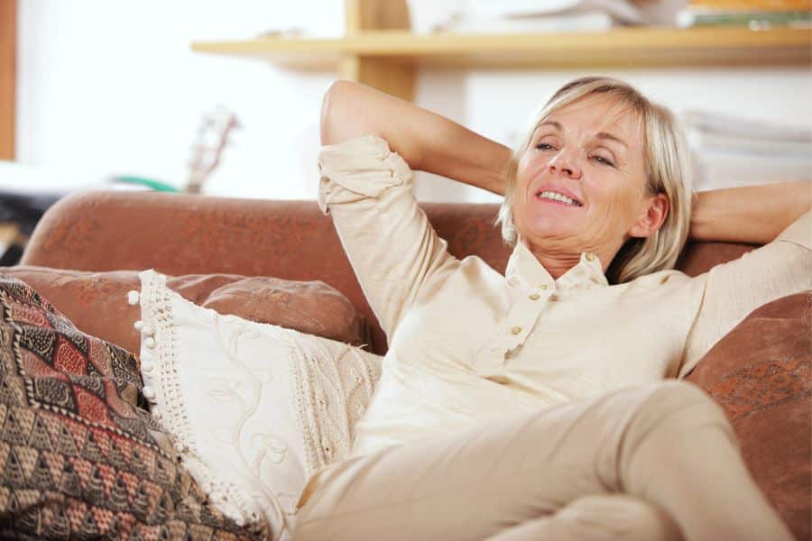 Relaxed older woman leaning back on a couch with her hands behind her head in a cozy living room. The scene conveys comfort and wellbeing after making healthy lifestyle changes.