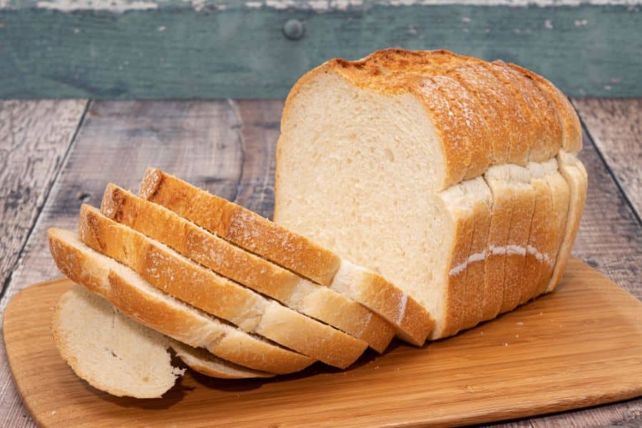 Loaf of white bread on a wooden cutting board with several slices cut and leaning forward. The plain sliced bread highlights a simple refined carbohydrate food.