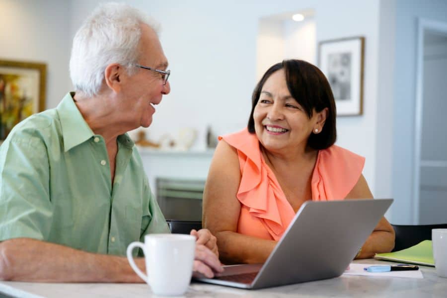 Two older adults smile at each other while sitting with a laptop and coffee mugs at a table. The image suggests a relaxed conversation about retirement plans or household finances.