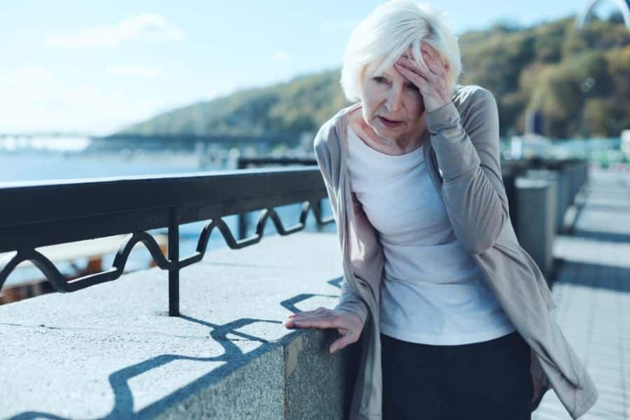 Elderly woman outdoors leans heavily against a waterfront railing with one hand on her forehead as if suddenly faint or disoriented. Her posture suggests dizziness or weakness that could be warning signs of stroke symptoms.