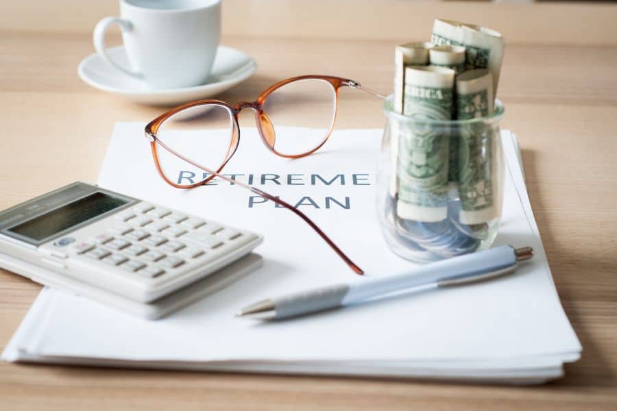 Eyeglasses calculator pen and a glass filled with rolled cash rest on top of papers labeled "RETIREMENT PLAN." The image shows financial planning materials arranged on a desk to represent retirement savings.