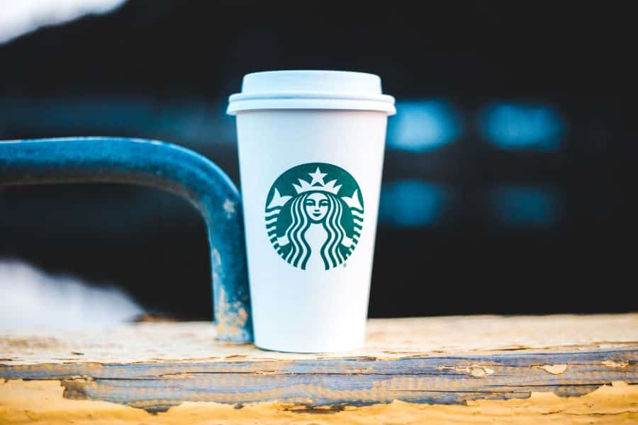 White Starbucks takeaway cup with the green logo sits on a weathered wooden railing against a dark blurred background. This supports content about save money on daily coffee purchases and small spending habits that add up.