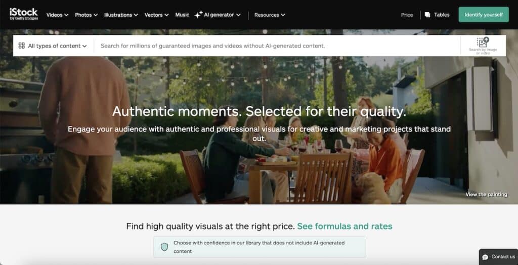 Screenshot of the iStock by Getty Images homepage with a blurred outdoor dinner scene in the hero banner and white navigation across the top. Visible text includes "iStock by Getty Images" "Videos" "Photos" "Illustrations" "Vectors" "Music" "AI generator" "Resources" "Price" "Tables" "Identify yourself" "All types of content" "Search for millions of guaranteed images and videos without AI generated content." "Search by image or video" "Authentic moments. Selected for their quality." "Engage your audience with authentic and professional visuals for creative and marketing projects that stand out." "View the painting" "Find high quality visuals at the right price. See formulas and rates" "Choose with confidence in our library that does not include AI generated content" and "Contact us."