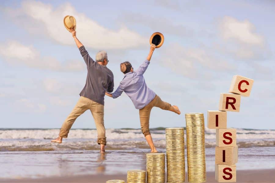 Older couple on a beach holding hands and lifting their hats as they balance playfully near stacked coins and wooden blocks spelling "CRISIS." The concept combines retirement lifestyle with financial strategies for handling market uncertainty.