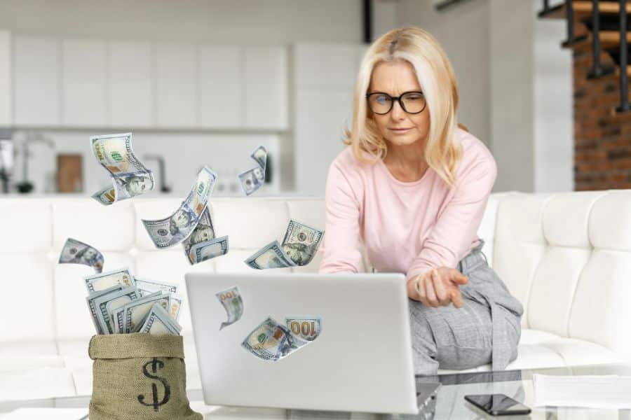 Blonde woman in glasses sits on a white couch working on a laptop while dollar bills appear to fly around her and a money bag sits on the table in front. The scene suggests an online business or financial opportunity being managed from home.
