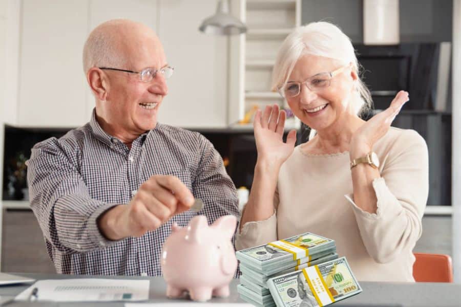 An older couple smiles at a kitchen table as the man drops coins into a pink piggy bank beside stacks of cash. This photo supports the idea of senior discounts by showing retirees saving money and feeling excited about stretching their budget.