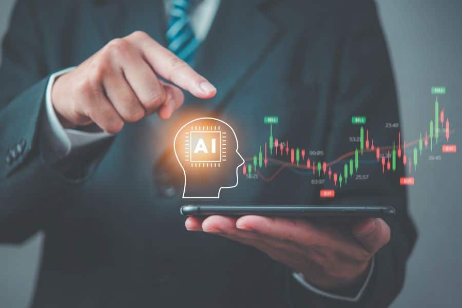 Business professional in a suit taps a tablet displaying a glowing AI head icon and a rising stock market chart with labels like "BUY" and "SELL". The scene represents using artificial intelligence to analyze trends and identify AI stocks to invest in.