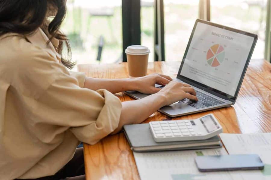 A woman works on a laptop at a wooden table with a calculator notebook phone and coffee nearby. The screen shows a budget chart which fits content about planning finances before investing in index funds.