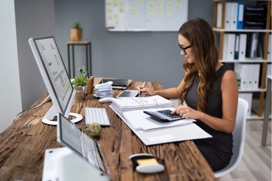 Woman at a desk reviews paperwork with a calculator and computer highlighting careful financial tracking and paying less tax through organized record keeping. Office setting with folders and notes shows detailed money management.