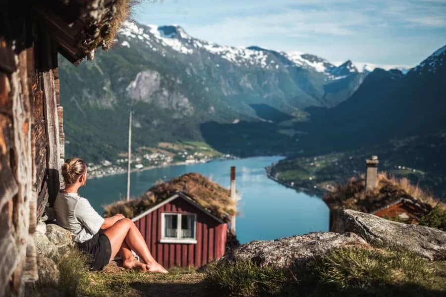 A woman sits outside a rustic cabin overlooking a deep blue fjord with snow capped mountains in the distance. This peaceful mountain view reflects Scandinavian lifestyle habits centered on quiet time in nature and simple surroundings.