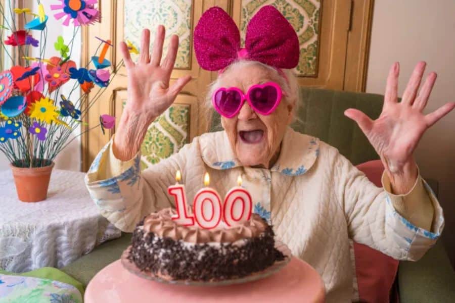 Elderly woman wearing bright pink heart shaped sunglasses and a large glittery bow throws both hands up in excitement behind a birthday cake with candles that read "100." The playful celebration fits a Centenarian Secrets theme by showing joy and personality at a 100th birthday.