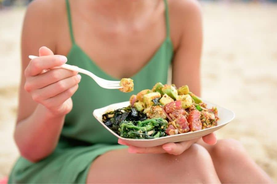 Person in a green dress sits on the beach holding a bowl of poke with tuna, avocado, seaweed, and greens while lifting a bite with a white plastic fork. This seafood meal works well for content about foods that stop memory loss because it shows a fresh dish with fish and nutrient rich ingredients.