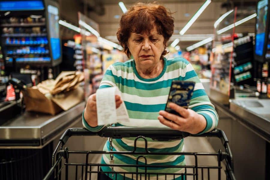 Older woman stands at a grocery checkout holding a receipt in one hand and her phone in the other while looking worried over her shopping cart. This works well for content about retailer tricks because it shows a shopper double checking prices and charges in store.