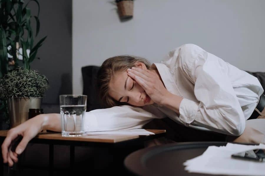 A woman in a white shirt rests her head on a desk while holding her temple, with a glass of water and papers nearby. Her posture and tired expression suggest fatigue or discomfort that can be linked to low in magnesium symptoms.