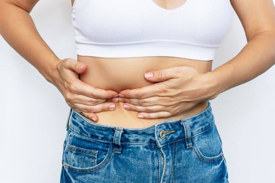 Person in a white sports bra and blue jeans presses both hands against the lower abdomen as if feeling pain or cramping. The close cropped view focuses on stomach discomfort that can appear alongside your liver related symptoms.