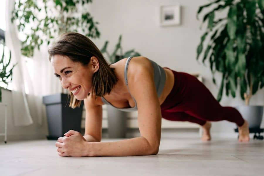 Smiling woman holds a forearm plank on a light wood floor with houseplants and soft window light in the background. The photo highlights core exercise as one way to support stronger posture habits at home.