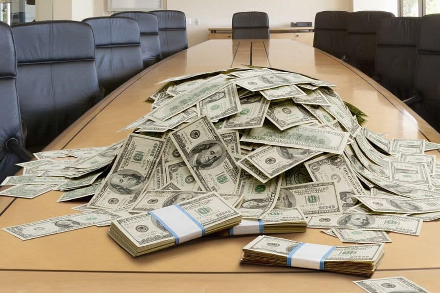 Piles of U.S. dollar bills and bundled stacks of cash are spread across a long conference table surrounded by black office chairs. It suggests a large amount of money in a business setting, which can support a point about retirement savings growth or financial risk.