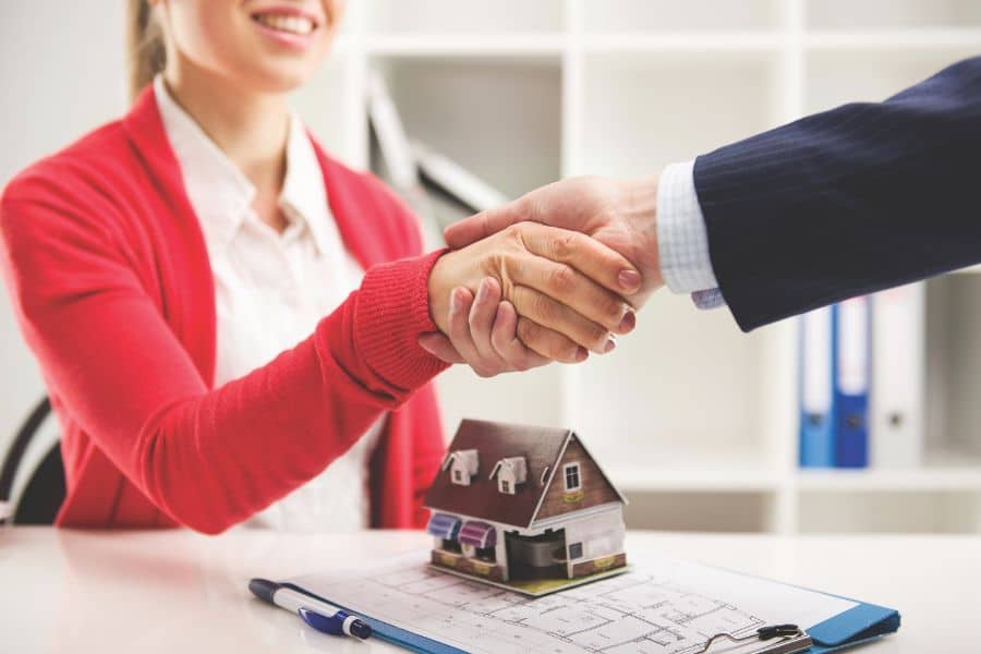 Two people shake hands across a desk with a small model house and paperwork in the foreground. The scene suggests a completed loan or financing agreement related to property.
