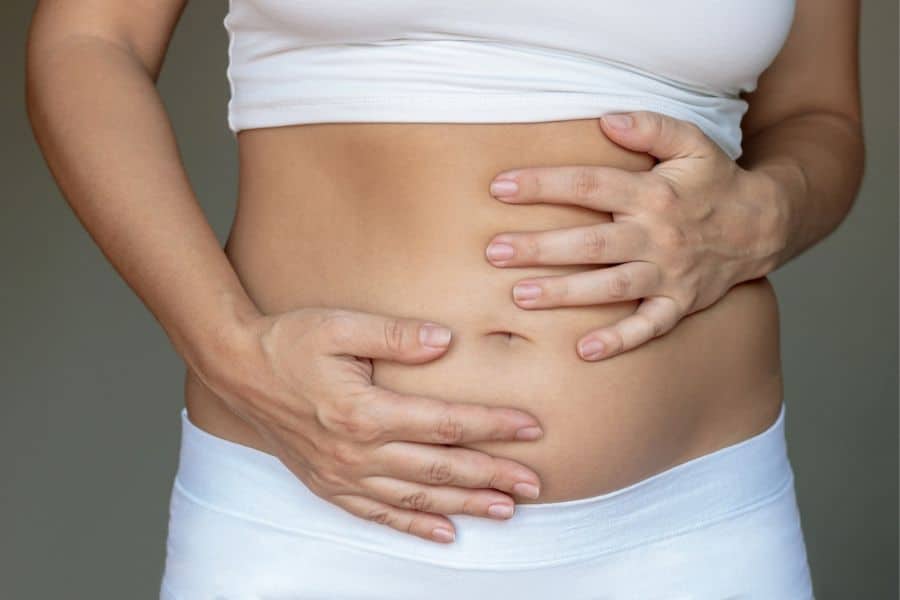 Cropped view of a person in white clothing holding both hands over their lower abdomen. The pose suggests stomach pain or discomfort.