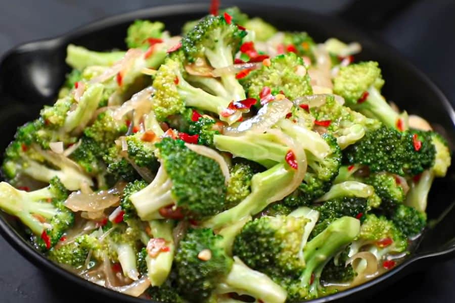 Black bowl filled with bright green broccoli florets tossed with sliced onions and red chili pieces in a glossy sauce. This savory vegetable dish fits content about foods that stop memory loss because it shows broccoli prepared in an appealing way for a brain healthy meal.