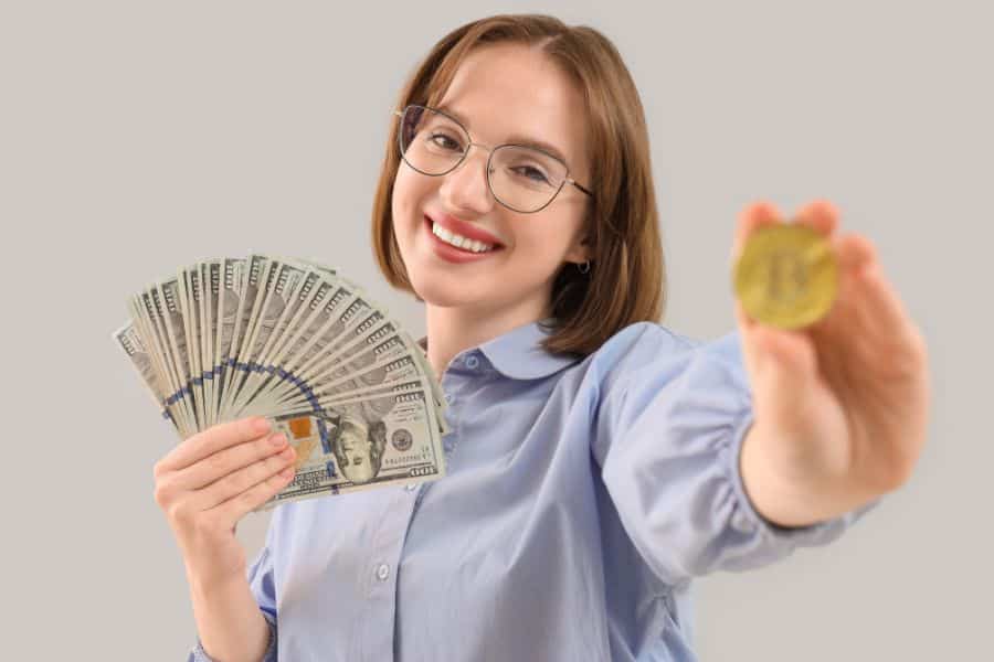 Smiling woman in glasses and a light blue shirt holds a fan of one hundred dollar bills while extending a gold coin toward the camera. The plain background keeps attention on the money and coin as symbols of savings or investing.