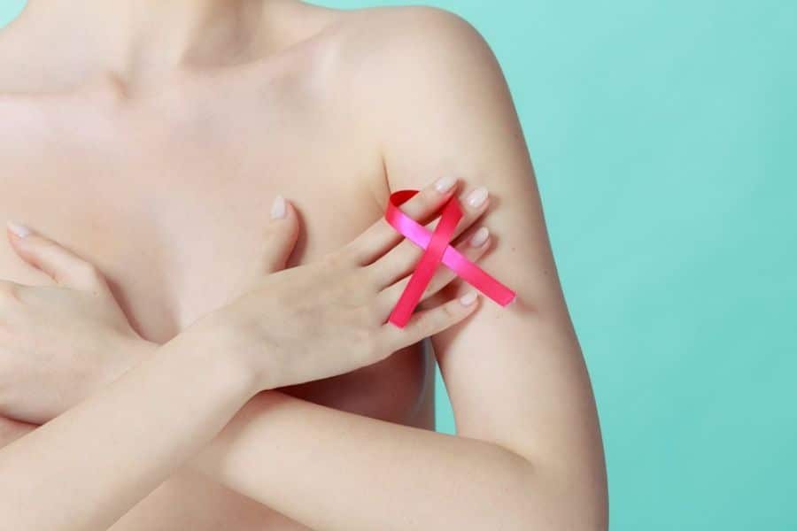 Bare upper body of a person covering their chest with one arm while holding a pink awareness ribbon against the shoulder. The photo represents breast cancer awareness and screening.