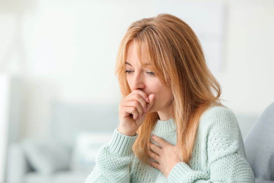 Woman in a light sweater coughs into her hand while the other hand rests on her chest. The scene suggests a respiratory symptom that may be discussed alongside early stages of cancer.