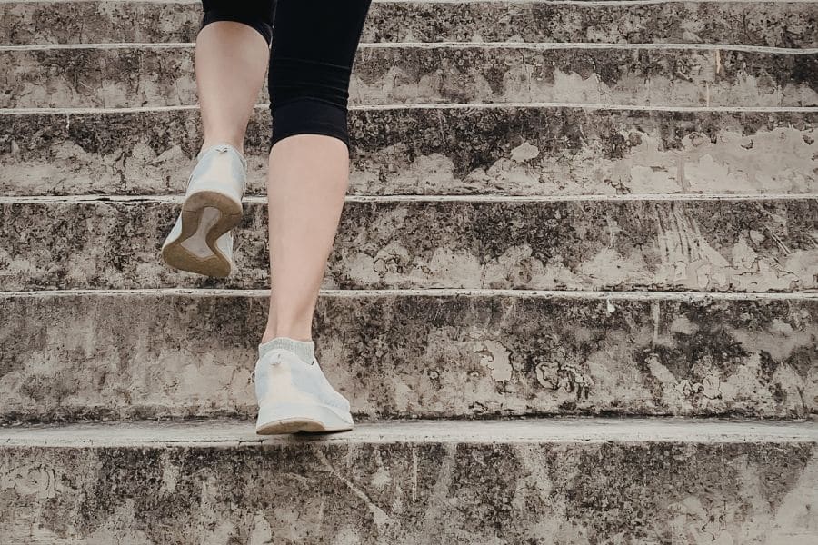 Close view of a person in white sneakers climbing a wide set of stone stairs. The upward movement supports content about mobility and daily activity as signs connected to how long you're going to live.