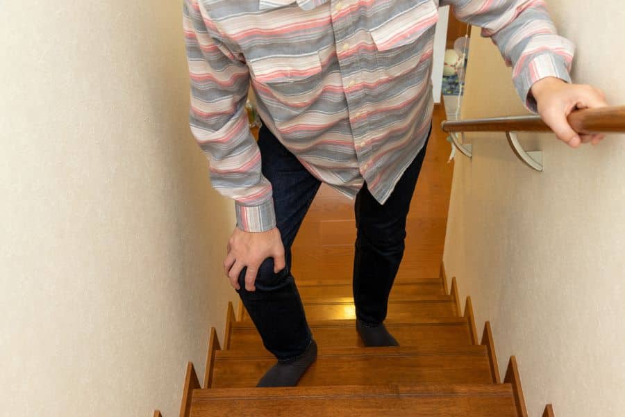 Person carefully climbs a narrow wooden staircase while holding the handrail and bracing one knee with the other hand. This highlights balance and physical strain in context about factors that may affect how long you're going to live.