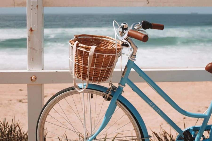 Light blue cruiser bicycle with a wicker basket is parked beside a beach with ocean waves in the background. This scene suggests gentle low impact movement that can pair well with joint strengthening exercises.
