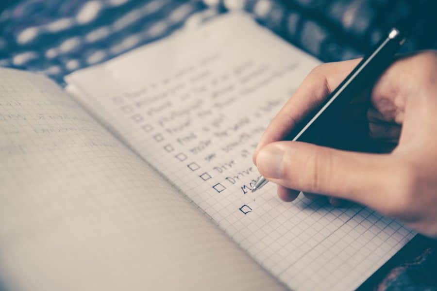Close up of a hand writing in a checklist notebook with several boxes marked and unmarked. The blurred list suggests bedtime planning or habit tracking related to managing lack of sleep.