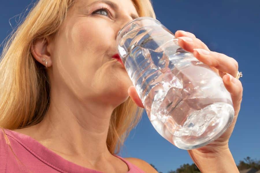 Close up of a woman drinking a large glass of water outdoors against a bright blue sky. The image supports hydration as a healthy habit in content about chronic inflammation.