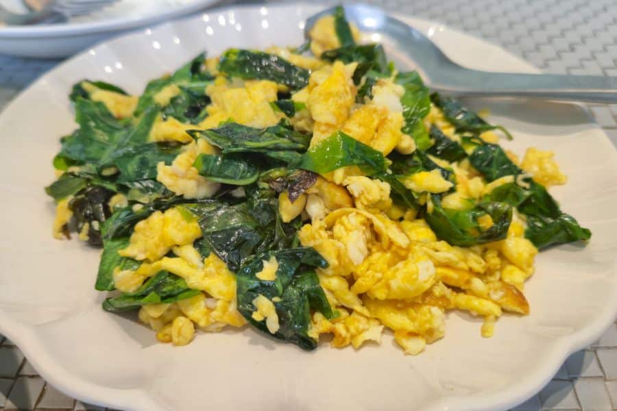 Plate of scrambled eggs mixed with leafy greens on a white dish with a spoon resting at the edge. The meal looks like a simple savory breakfast or light lunch.