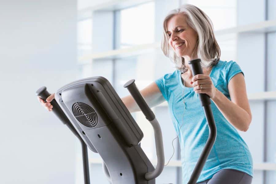 Smiling older woman exercises on an elliptical machine indoors while wearing earphones and a blue shirt. The photo shows a cardio activity often used alongside joint strengthening exercises to support mobility and endurance.